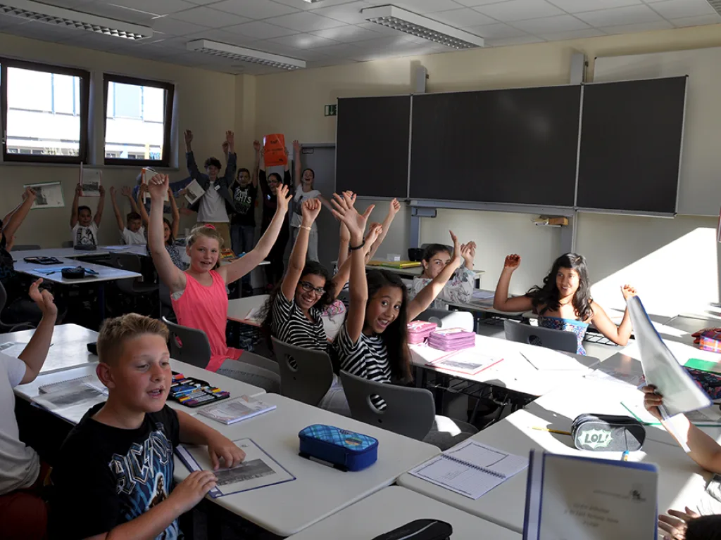 Kinder heben am Tisch beide Arme und halten Blätter hoch.